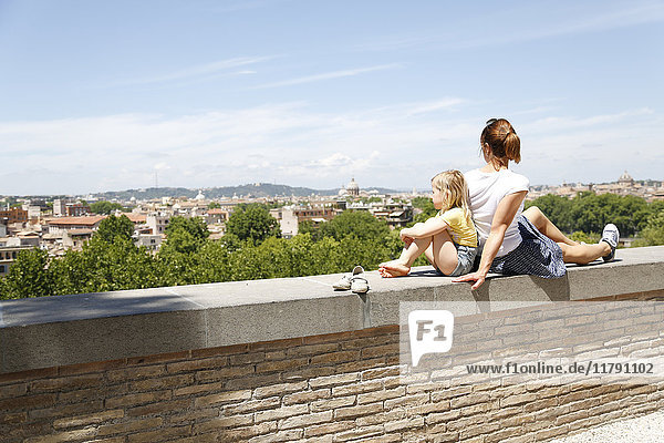 Italy  Rome  mother and her little daughter sitting back to back on a wall looking at view
