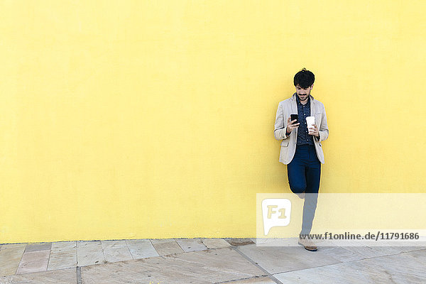 Junger Mann mit Kaffee  um das Handy vor der gelben Wand zu überprüfen.