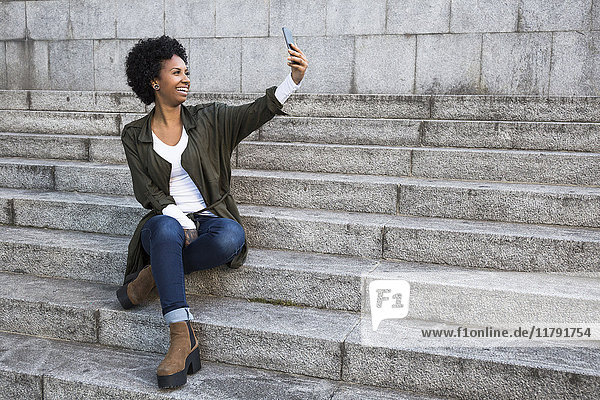 Lächelnde junge Frau sitzt auf einer Treppe und nimmt Selfie mit dem Smartphone.