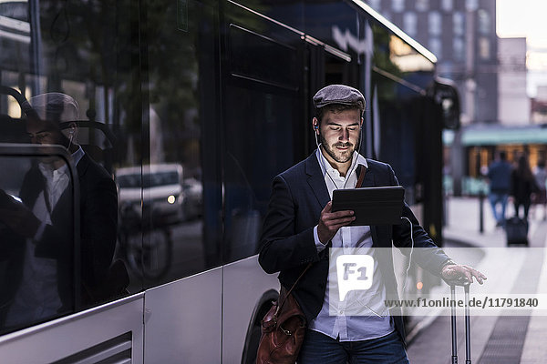 Junger Mann an der Bushaltestelle in der Stadt mit Kopfhörer und Tablett