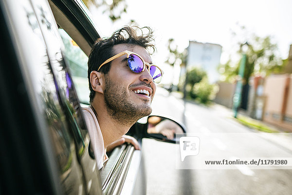 Mann mit Sonnenbrille  der sich aus dem Fenster eines fahrenden Autos lehnt.