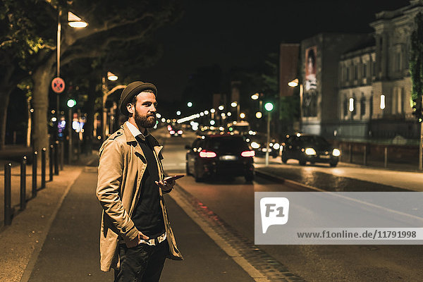 Stilvoller junger Mann mit Handy auf urbaner Straße bei Nacht