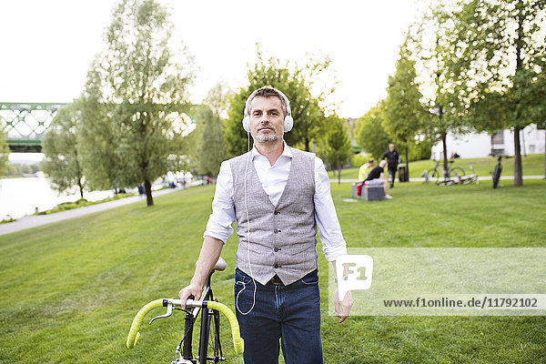 Selbstbewusster Geschäftsmann mit Fahrrad und Kopfhörer im Stadtpark