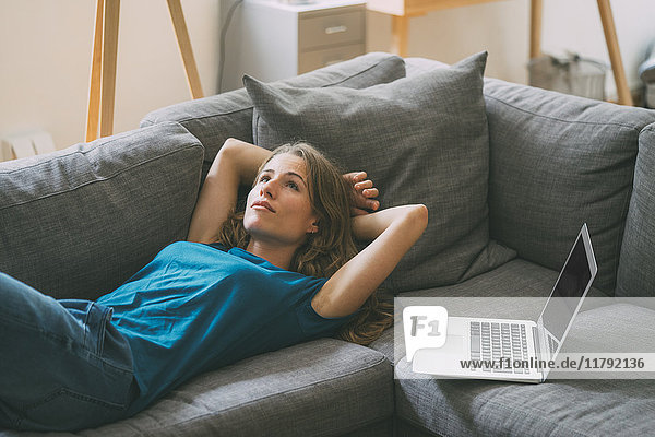 Junge Frau mit Laptop zu Hause auf der Couch liegend