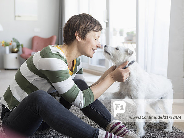Lächelnde Frau sitzt auf dem Boden im Wohnzimmer und kuschelt mit ihrem Hund.