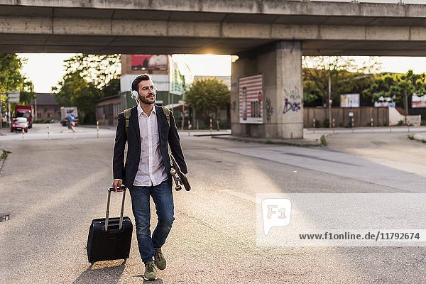 Young man on the move with skateboard  rolling suitcase and headphones