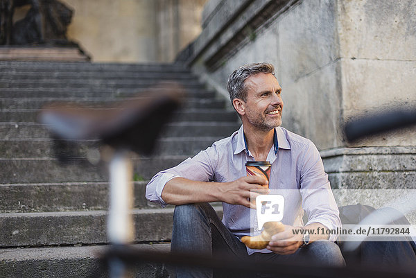 Lächelnder Mann sitzt auf einer Treppe mit Croissant und Kaffee zum Mitnehmen in der Stadt.
