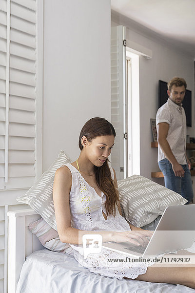 Young woman using laptop in bedroom with man in background