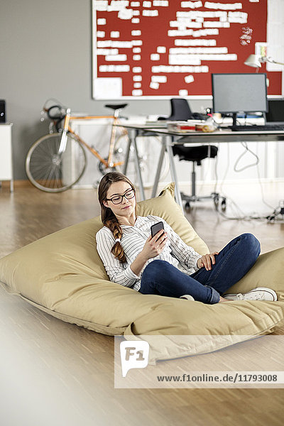 Young woman with cell phone sitting in bean bag in office