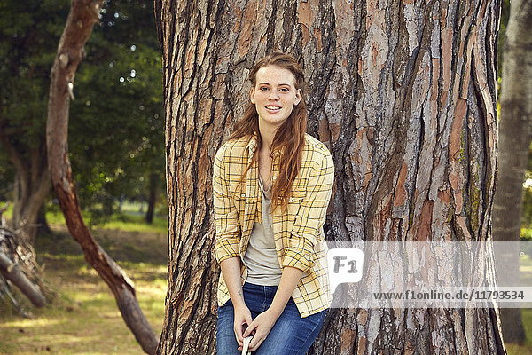 Porträt einer lächelnden jungen Frau  die sich an einen Baumstamm lehnt.
