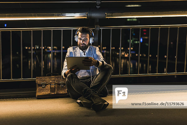 Smiling young man with tablet and headphones sitting on bridge at night