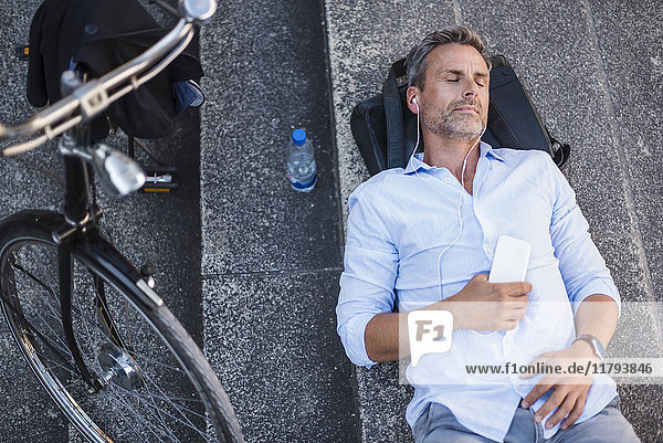 Mann mit geschlossenen Augen auf Treppe liegend mit Handy und Ohrstöpseln neben Fahrrad