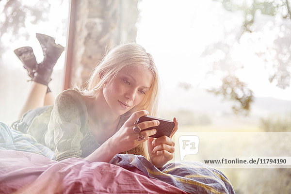 Porträt einer jungen Frau auf dem Bett liegend mit Blick auf das Handy