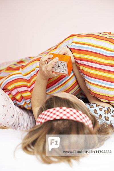 Rückansicht des kleinen Mädchens das im Bett liegt und den Wecker stellt.