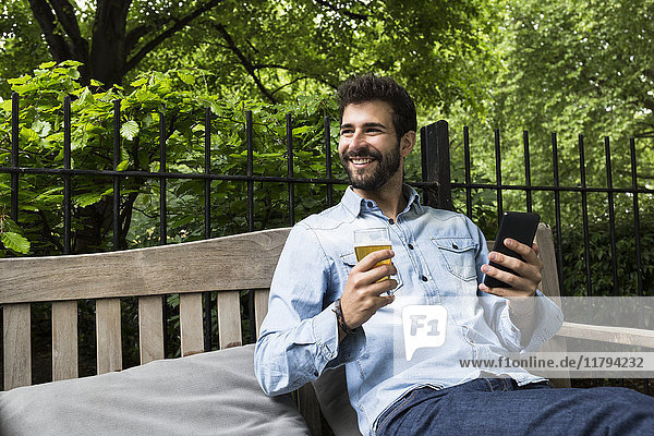 Porträt eines lächelnden jungen Mannes mit Handy und einem Glas Bier im Garten