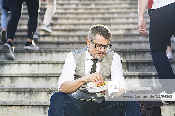 Geschäftsmann in der Stadt sitzt auf einer Treppe und isst einen Hamburger.