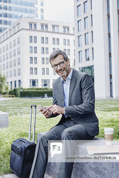 Smiling mature businessman outdoors with laptop  cell phone  takeaway coffee and rolling suitcase