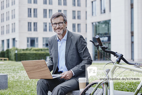 Portrait of smiling mature businessman outdoors with laptop  takeaway coffee and bicycle