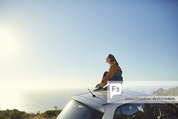 Südafrika  Kapstadt  Signal Hill  junge Frau sitzt auf dem Auto und genießt den Blick aufs Meer.