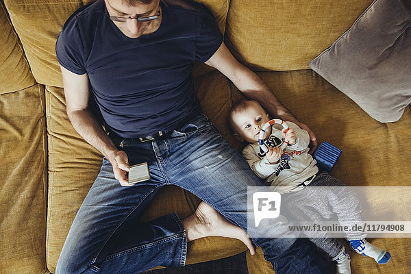 Kleiner Junge  der neben seinem Vater auf der Couch liegt und ein Smartphone benutzt.