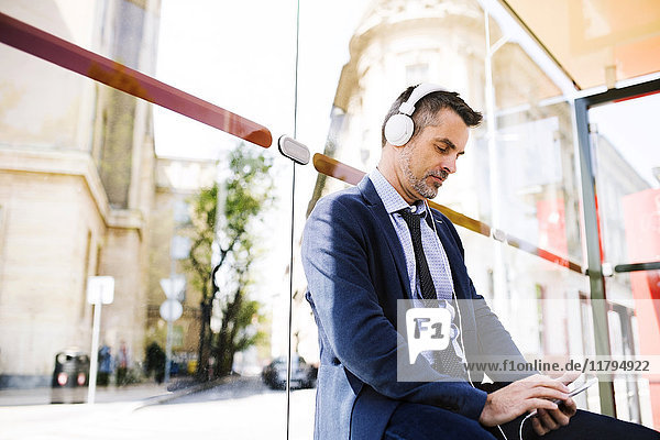 Geschäftsmann mit Smartphone und Kopfhörer wartet an der Bushaltestelle