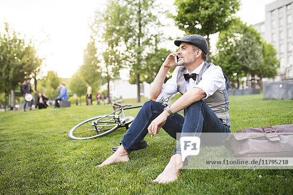Confident mature businessman with bicycle and smartphone in the city park sitting on grass