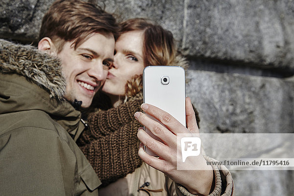 Glückliches junges Paar  das sich im Freien küsst und einen Selfie nimmt.