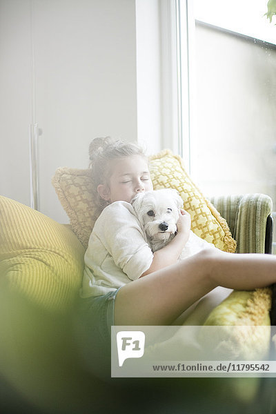 Little girl sitting on armchair at home cuddling with her dog