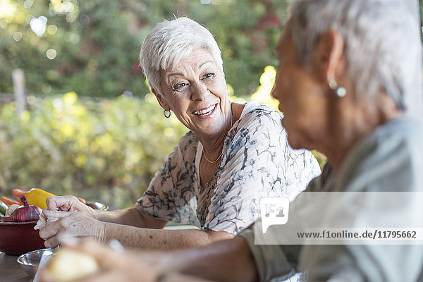 Porträt einer lächelnden Seniorin  die ihrem Freund auf der Terrasse beim Zubereiten des Mittagessens zuhört.
