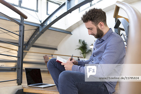 Der Mann im modernen Büro sitzt auf einer Treppe und macht sich Notizen.