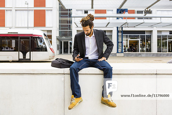 Junger Geschäftsmann mit Dreadlocks mit Smartphone beim Warten am Bahnhof