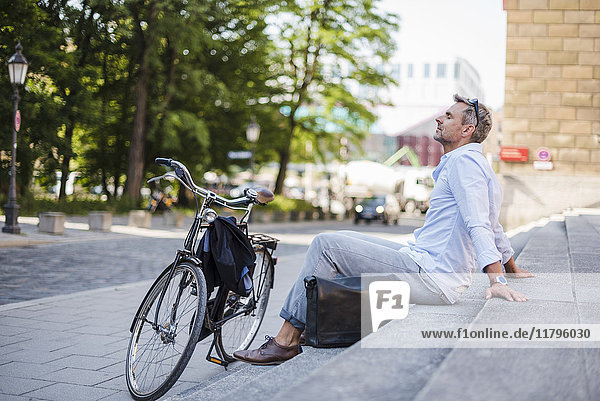 Entspannter Mann sitzt auf einer Treppe in der Stadt neben dem Fahrrad.