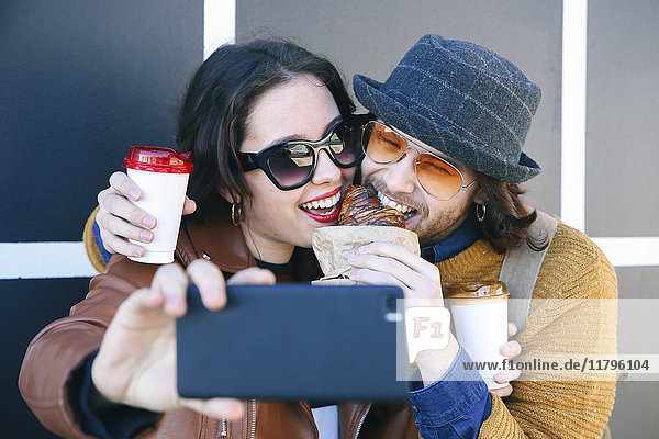 Young couple with croissant and coffee to go taking selfie with smartphone