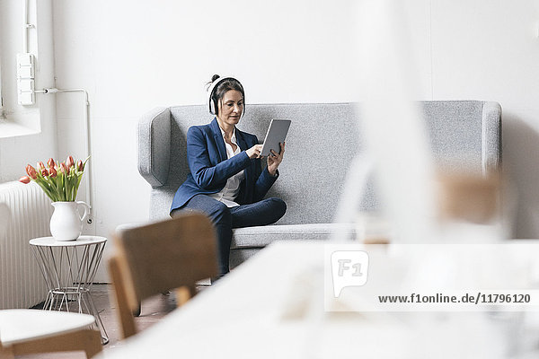 Businesswoman sitting on couch in a loft using tablet and headphones