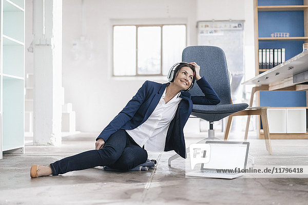 Smiling businesswoman sitting on the floor in a loft listening music with headphones