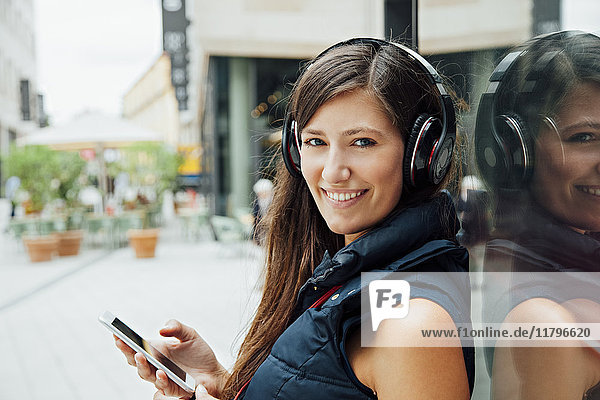 Porträt der lächelnden jungen Frau mit Kopfhörer und Handy in der Stadt
