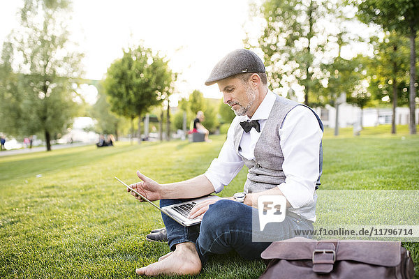 Selbstbewusster reifer Geschäftsmann mit Laptop im Stadtpark auf Rasen sitzend