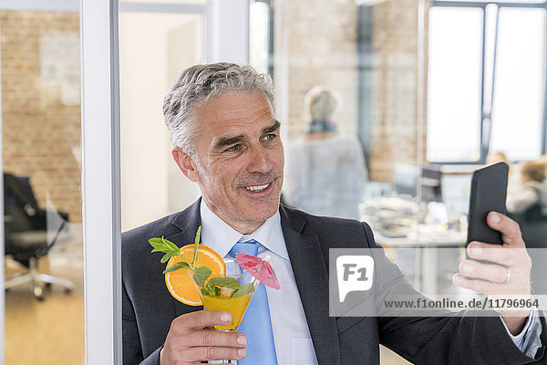 Ein reifer Geschäftsmann trinkt einen Cocktail  während er einen Selfie nimmt.