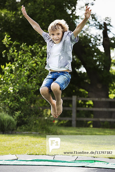 Boy wearing shirt and denim shorts jumping on a trampoline in a garden.