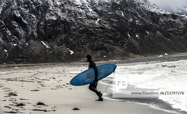 Ein Surfer  der einen Neoprenanzug trägt und ein Surfbrett mit Bergen im Rücken aus dem Meer trägt.