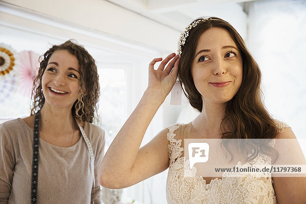 Eine junge Frau probiert Haarschmuck und Schleier mit weißer Spitze über dem Brautkleid an  unterstützt von einer Einzelhandelsberaterin.
