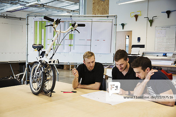 Drei Männer bei einer Besprechung in einer Fahrradfabrik  an einem Tisch sitzend mit einem Klappfahrrad auf der Tischplatte.