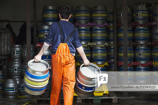 Rear view of man working in a brewery carrying a metal beer keg in either hand.