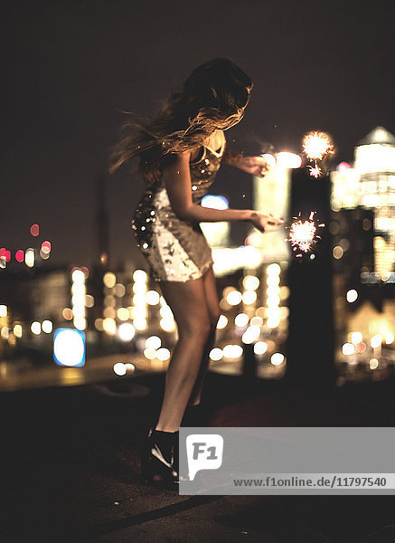 A young woman holding a sparkler and dancing on a building rooftop at night