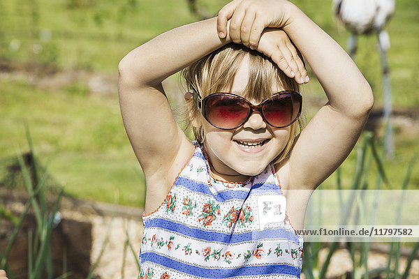 Girl in a sundress wearing sunglasses  hands on head  smiling at camera.