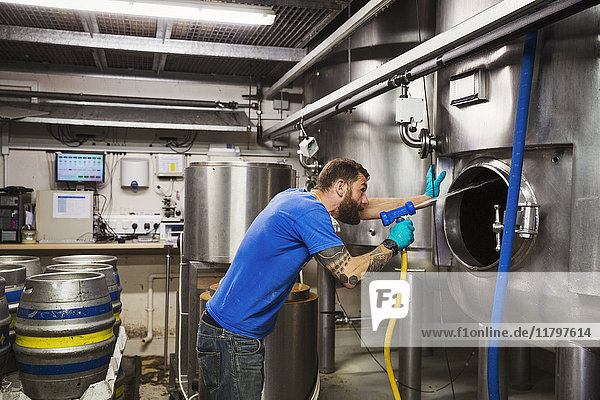 Mann  der in einer Brauerei arbeitet und das Innere eines großen Edelstahlkessels mit einem Hochdruckreiniger reinigt.