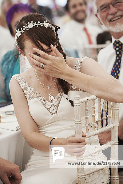 Braut im Brautkleid in einem Festzelt sitzend  lachend und ihr Gesicht bedeckend.
