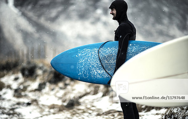 Ein Surfer  der einen Neoprenanzug trägt und Surfbretter trägt  steht an einem verschneiten Strand mit Bergen im Rücken.