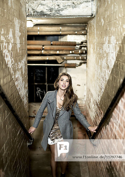 A young woman in a dress and jacket climbing up a narrow stairway in a building. Pipework and brick work.