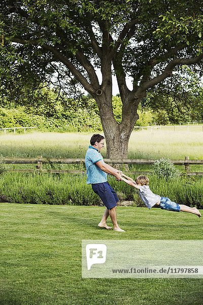 Man standing on a lawn in a garden  holding a boy by his hands  spinning and whirling him around.